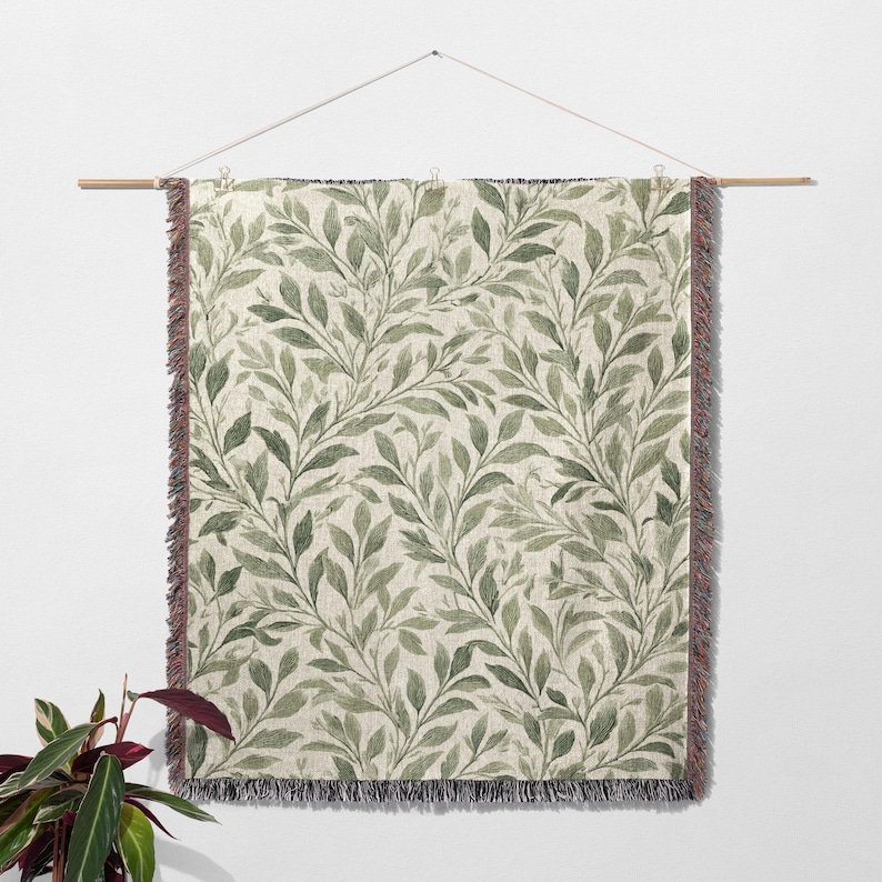 Op de afbeelding: Een rechthoekige wandhanger met een botanisch bladerpatroon in groen en beige. De bladeren zijn gerangschikt in een vloeiend, naturalistisch ontwerp. De randen hebben een gefranjerde rand. De wandhanger is opgehangen aan een houten roede.