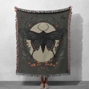 May include: A dark green woven throw blanket featuring a black and orange butterfly design. The butterfly is encircled by a wreath of leaves and berries.