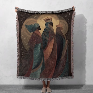 May include: A woven throw blanket depicting the Three Wise Men. The design features figures in robes and crowns, set against a golden halo and stars. The blanket has a fringed edge and a colour palette of browns, blues, and golds. The blanket is approximately 152 cm x 127 cm.