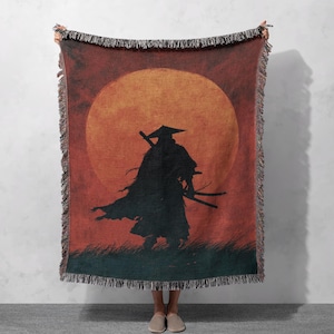 May include: A woven throw blanket featuring a silhouette of a samurai warrior with two swords against a large orange sun. The blanket has a fringed edge and a dark green base with grass detail.