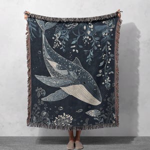 May include: A woven throw blanket with a blue background and a white whale design. The whale is surrounded by white floral designs and bubbles. The blanket has a fringe border.