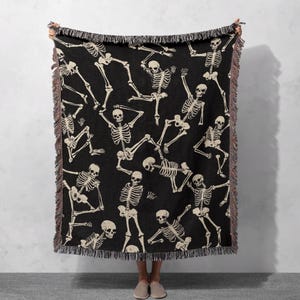 May include: Black woven throw blanket with a repeating pattern of white skeletons in various poses.