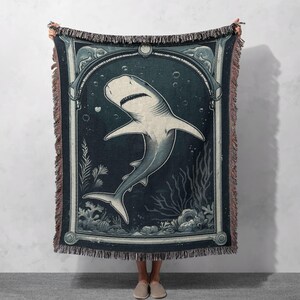 May include: A woven tapestry throw blanket featuring a vintage illustration of a shark swimming in the ocean. The shark is white with a dark blue outline and is surrounded by seaweed and bubbles. The background is a dark blue color with a light blue border.