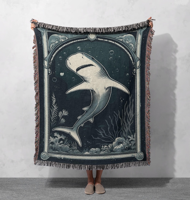 May include: A woven tapestry throw blanket featuring a vintage illustration of a shark swimming in the ocean. The shark is white with a dark blue outline and is surrounded by seaweed and bubbles. The background is a dark blue color with a light blue border.