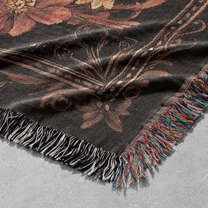 May include: A black woven throw blanket with a floral pattern in shades of brown and gold. The blanket has a fringe border.