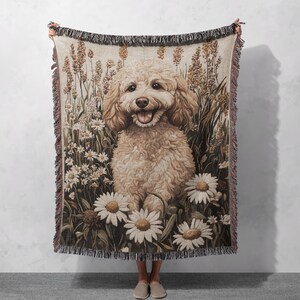 May include: A woven tapestry throw blanket featuring a brown and white poodle sitting in a field of daisies. The dog is smiling and has a happy expression.