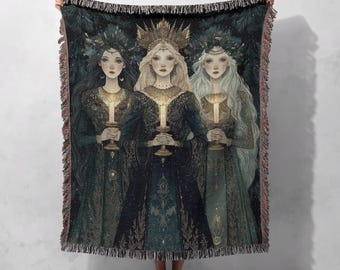 May include: A woven throw blanket featuring a detailed illustration of three women holding candles. The women are adorned in ornate gowns and headdresses, with long hair. The color palette includes shades of green, blue, and gold, with a dark background.