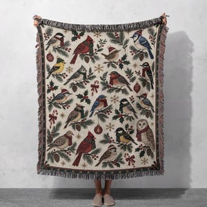 Könnte beinhalten: Eine gewebte Wohndecke mit cremefarbenem Hintergrund und einem wiederkehrenden Muster aus verschiedenen Vögeln mit Winterschals und Mützen, durchsetzt mit Tannenzweigen, Stechpalmenbeeren, Zuckerstangen, Weihnachtskugeln und Schneeflocken, mit Fransen.