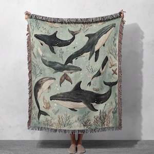 May include: A woven throw blanket with a blue background featuring a pattern of black whales and coral. The whales are depicted in various sizes and positions, swimming through the ocean.