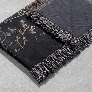 May include: A black woven throw blanket with a floral pattern in gold and white. The blanket has a fringe border.