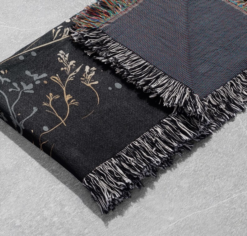 May include: A black woven throw blanket with a floral pattern in gold and white. The blanket has a fringe border.