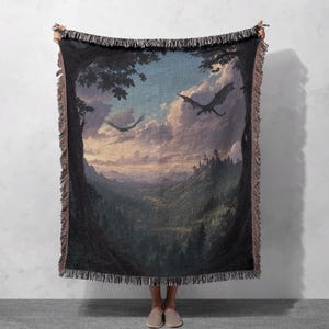 Può includere: Una coperta tessuta raffigurante un paesaggio fantasy con draghi che volano su una valle, un castello e una foresta. La tavolozza dei colori comprende sfumature di blu, verde e marrone. La coperta ha un bordo sfrangiato.