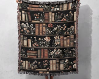 Può includere: Una coperta nera con un design a libreria, che mostra file di libri e composizioni floreali. I libri sono in varie tonalità di marrone e beige, con fiori bianchi, pesca e arancioni. La coperta ha un bordo sfrangiato.