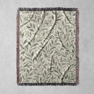 Op de afbeelding: Een rechthoekige geweven sprei met een botanisch bladerpatroon. De bladeren zijn in tinten groen en grijs op een cr&egrave;mekleurige achtergrond. De deken heeft een gefranjerde rand in zwart, rood en beige.