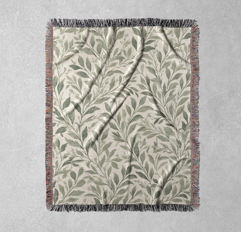 Op de afbeelding: Een rechthoekige geweven sprei met een botanisch bladerpatroon. De bladeren zijn in tinten groen en grijs op een cr&egrave;mekleurige achtergrond. De deken heeft een gefranjerde rand in zwart, rood en beige.