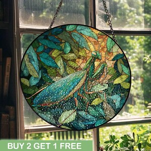 May include: Round stained glass window art featuring a praying mantis in a lush green and blue leaf setting. The insect is detailed with shades of blue and green. The artwork is suspended by a chain. The text "BUY 2 GET 1 FREE" is at the bottom.