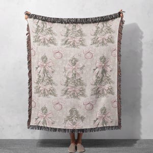May include: A woven throw blanket with a cream background and a repeating pattern of pink and green Christmas trees with pink bows and ornaments.
