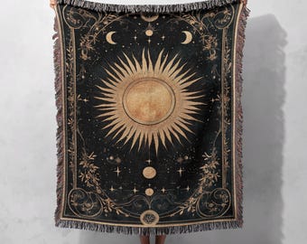 Può includere: Una coperta in tappezzeria nera e dorata con un design celeste che presenta un sole, una luna e stelle. La coperta ha un bordo a frangia.