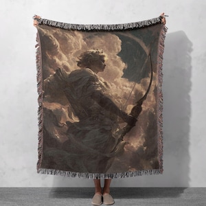 May include: A woven throw blanket featuring a classical painting of a figure with a bow and arrow against a cloudy backdrop. The blanket has a fringed edge and is primarily in shades of brown, beige, and gray.