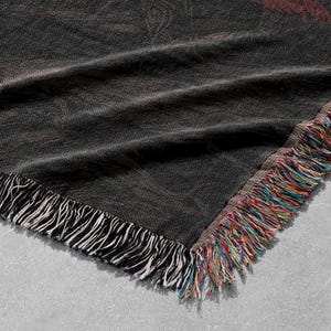 May include: A dark gray woven throw blanket with a fringed edge. The blanket has a textured appearance with a subtle pattern. The fringe is a mix of white, red, blue, and yellow threads. The blanket is folded.