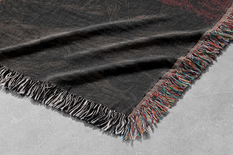 May include: A dark gray woven throw blanket with a fringed edge. The blanket has a textured appearance with a subtle pattern. The fringe is a mix of white, red, blue, and yellow threads. The blanket is folded.