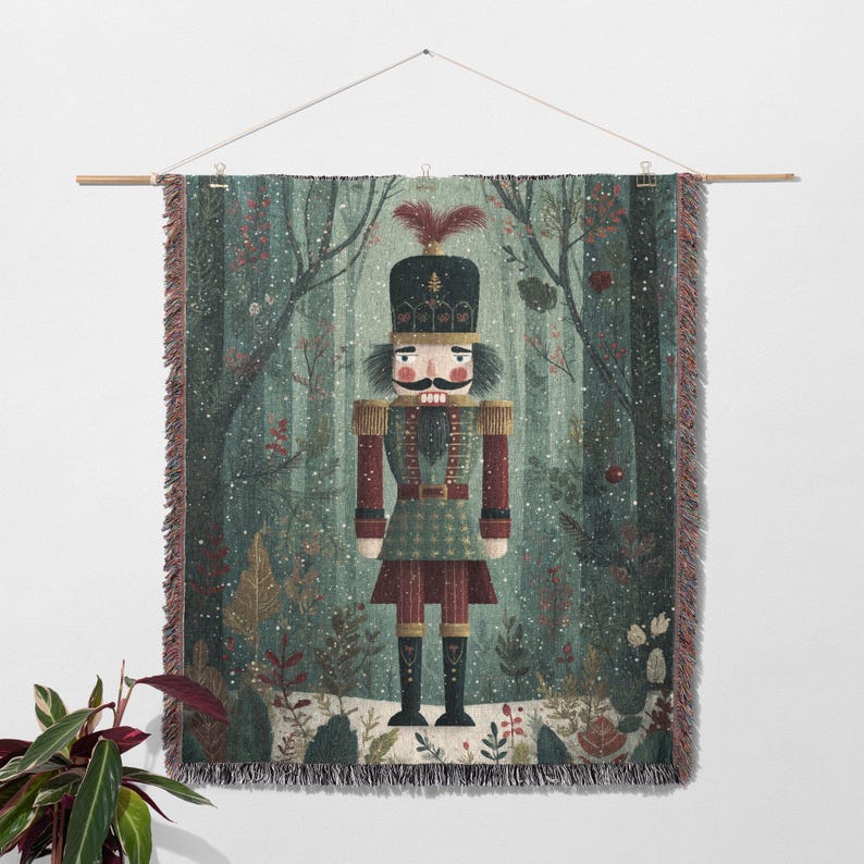 May include: A woven wall hanging featuring a detailed illustration of a nutcracker soldier in a forest setting. The nutcracker is dressed in a green and red uniform, with a black hat and a large mustache. The background depicts a snowy forest with trees and foliage.