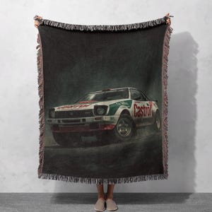 May include: A woven throw blanket depicting a vintage rally car in white, green, and red, with the word "Castrol" on the side. The blanket has a dark background and a fringed edge.
