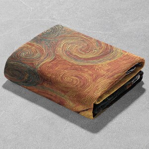 May include: Folded blanket featuring a swirling pattern in shades of orange, yellow, and teal. The design resembles brushstrokes or a stylized representation of a natural phenomenon. The blanket is folded neatly, showcasing the detailed texture and color variations.