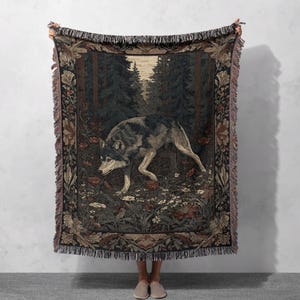 Peut inclure: Un plaid tissé présentant une illustration centrale d'un loup gris et noir marchant dans une forêt avec des pins sombres et de petites fleurs. La couverture possède une bordure florale décorative et des franges sur le haut et le bas.