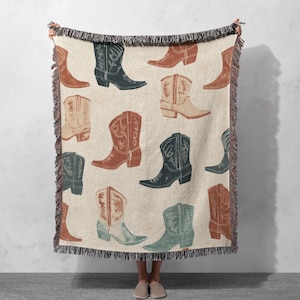 May include: A woven throw blanket with a pattern of cowboy boots in shades of brown, green, and blue. The boots are arranged in a repeating pattern on a cream-colored background.