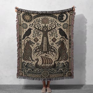 Peut inclure: Un plaid tissé présentant un motif central d'arbre, deux corbeaux, deux loups et un navire viking naviguant sur des vagues. Cette tapisserie beige et brun foncé comprend des motifs célestes de soleil et de lune, des runes et des franges.