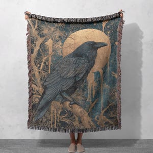 Può includere: Una coperta tessuta con una dettagliata illustrazione di un corvo appollaiato su un ramo contro uno sfondo di luna dorata e accenti verde acqua e oro. La coperta ha un bordo sfrangiato.