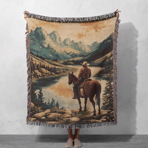 May include: A woven tapestry throw blanket featuring a vintage illustration of a cowboy riding a horse through a mountain landscape. The scene is set against a backdrop of a lake and lush green trees. The colours are muted and earthy, with shades of brown, green, and blue.