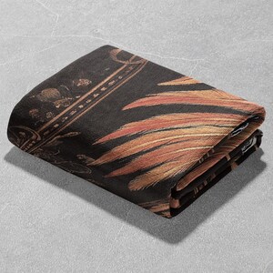 May include: A black and gold throw blanket with a feather pattern. The blanket is folded in half and has a textured surface.