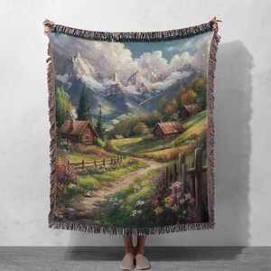 Puede incluir: Una manta de tejido de tapiz que presenta un paisaje pintoresco con un camino sinuoso que conduce a través de un valle verde exuberante hacia un grupo de encantadoras cabañas de madera ubicadas entre las laderas de montañas cubiertas de nieve. El cielo es de un azul vibrante con nubes blancas esponjosas.