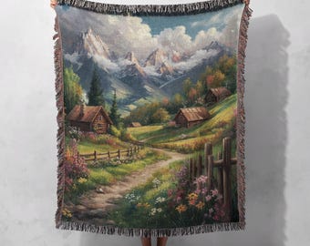 Può includere: Una coperta in tessuto di arazzo che presenta un paesaggio pittoresco con un sentiero tortuoso che attraversa una valle verde lussureggiante verso un gruppo di incantevoli baite in legno annidati tra le pendici di montagne innevate. Il cielo è di un blu vibrante con nuvole bianche soffici.
