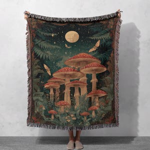 May include: A woven throw blanket featuring a whimsical forest scene with large red and white spotted mushrooms under a full moon and starry sky. The blanket has a dark green and brown color palette with fringed edges.