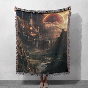 May include: Woven throw blanket featuring a fantasy landscape with a city, mountains, and a large planet in the sky. The color palette includes shades of brown, orange, and green. The blanket has a fringed edge.