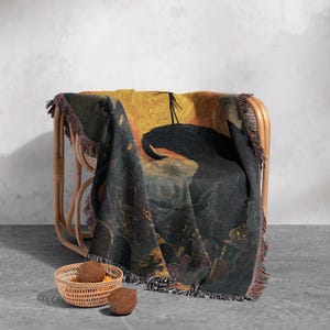 May include: A woven Halloween-themed throw blanket draped over a wicker chair. The blanket features a dark, detailed illustration with a black silhouette against a yellow and orange background. The blanket has a fringed edge and is paired with a small basket and two coconuts.