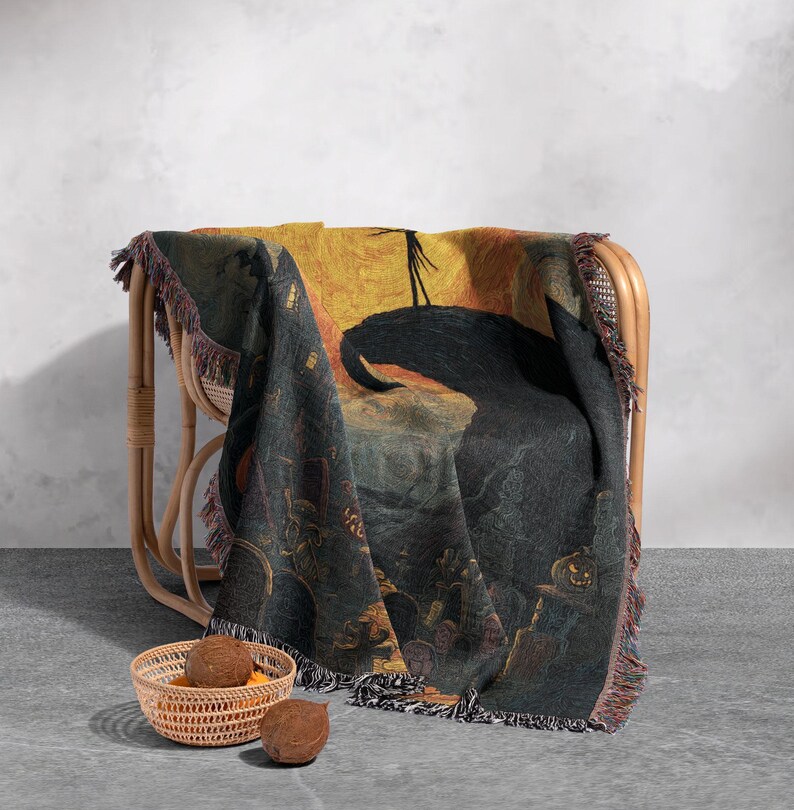 May include: A woven Halloween-themed throw blanket draped over a wicker chair. The blanket features a dark, detailed illustration with a black silhouette against a yellow and orange background. The blanket has a fringed edge and is paired with a small basket and two coconuts.