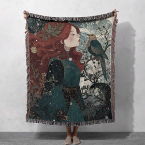 May include: A woven throw blanket featuring an illustration of a woman with flowing red hair and a teal dress, with a bird perched on a branch. The blanket has a fringed edge and a color palette of teal, red, and cream.