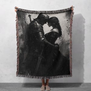 Op de afbeelding: Een zwart-wit geweven wandkleed met een ridder en een vrouw die elkaar omhelzen. De ridder draagt ​​een harnas en houdt een zwaard vast. De vrouw draagt ​​een lange jurk. De achtergrond is een donker, getextureerd oppervlak.