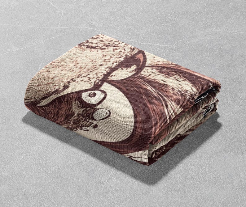 May include: A folded brown and beige throw blanket with a graphic print of a cartoon animal face with large eyes.