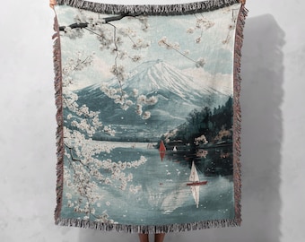 May include: Woven throw blanket depicting a mountain landscape with sailboats and cherry blossoms. The blanket has a fringed edge and a colour palette of blues, greens, and white. The scene is reminiscent of a Japanese painting.