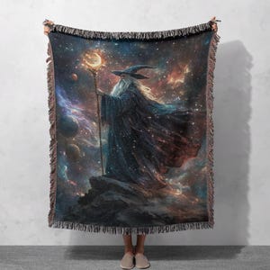 Puede incluir: Una manta tejida que representa a un mago sosteniendo un bastón con una luna creciente brillante. El mago se encuentra sobre un fondo de una galaxia estrellada con planetas y nebulosas. La manta tiene un borde con flecos.