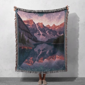 May include: Woven throw blanket featuring a scenic mountain and lake landscape. The design showcases a mountain range with snow-capped peaks reflected in the calm water. The blanket has a fringed edge and a decorative border, with colors of purple, orange, and green.