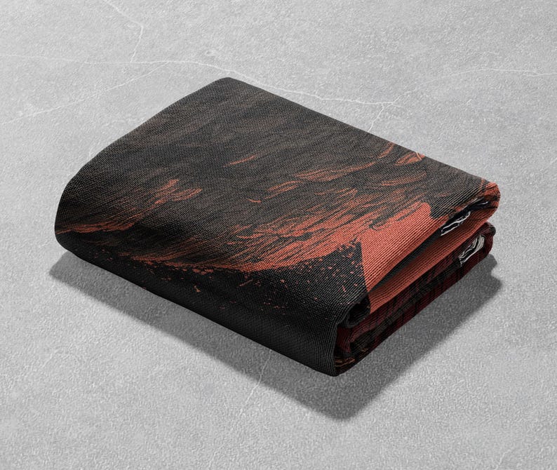 May include: Folded rectangular textile with a dark brown and red abstract design. The fabric appears to be a woven material, with the design visible on the top layer and the edges. The folded edges reveal a contrasting red color.