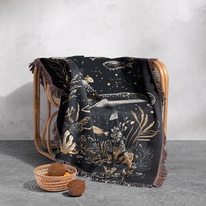 May include: A black woven throw blanket with a gold and white design of fish and seaweed. The blanket is draped over a wicker chair and has fringe on the edges.
