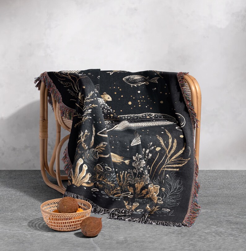 May include: A black woven throw blanket with a gold and white design of fish and seaweed. The blanket is draped over a wicker chair and has fringe on the edges.