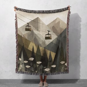 May include: A woven throw blanket featuring a mountain landscape with two cable cars suspended on wires above a forest of dark green pine trees and a foreground of white wildflowers. The blanket has a fringed edge and a muted color palette.
